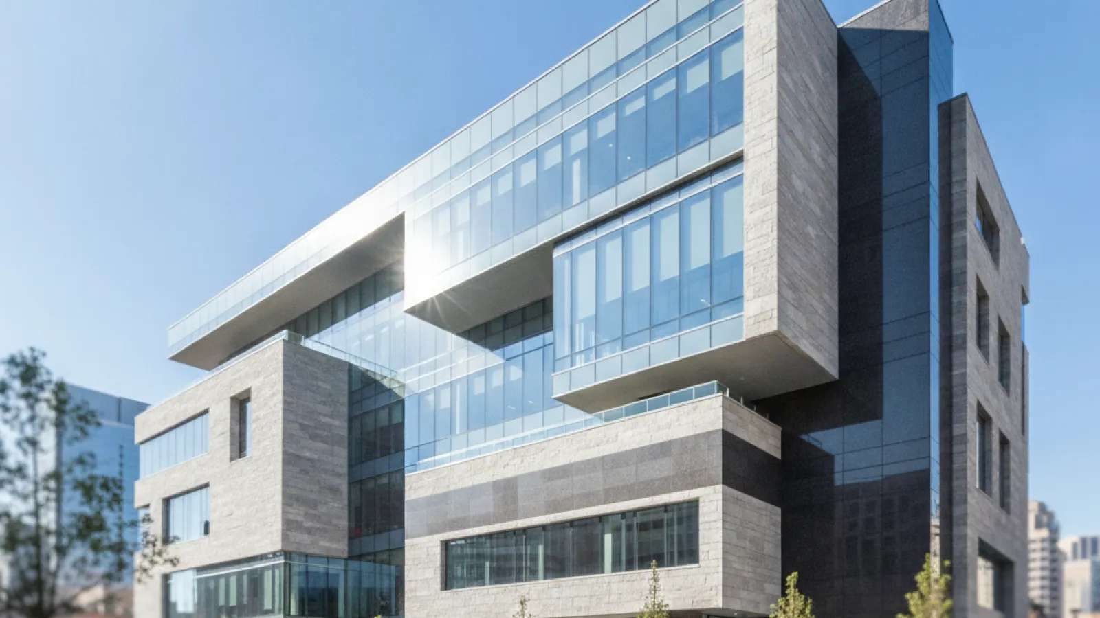 A modern building with reflective glass and textured stone facades stands under a clear, sunny sky, fronting a paved plaza with trees in planters and geometric concrete benches.