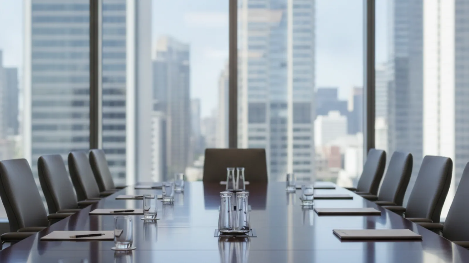 An empty, naturally lit boardroom features a long, polished wooden table set with glasses and papers, surrounded by office chairs, with city skyscrapers visible through large windows.