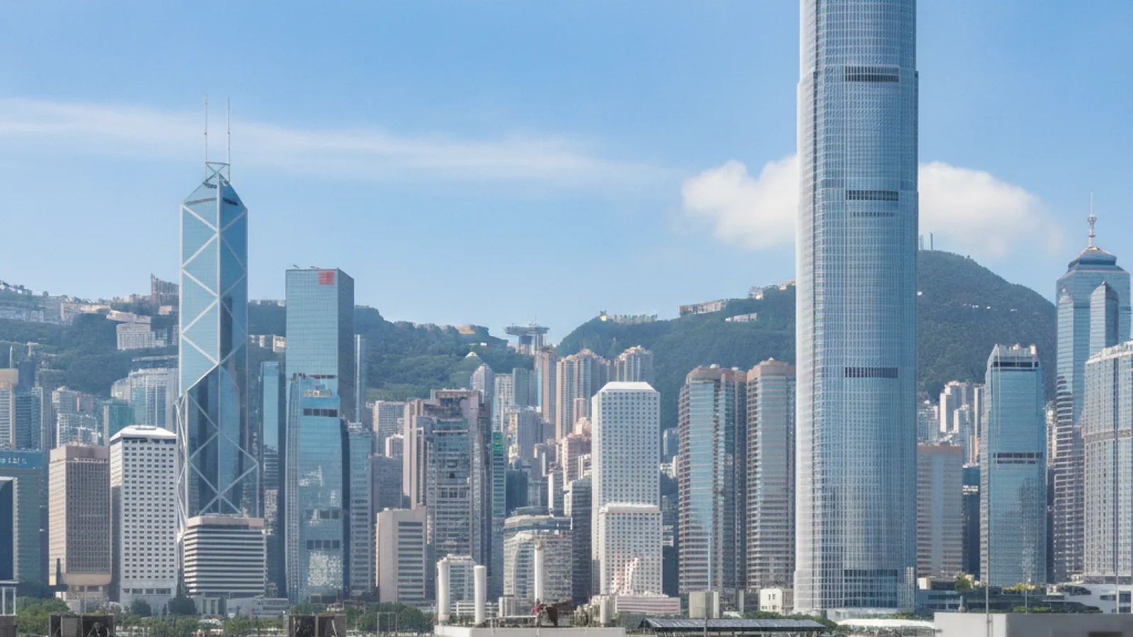 On a sunny day, Hong Kong's urban skyline features numerous skyscrapers, including the International Finance Centre and Bank of China Tower, backed by green hills under a bright blue sky with white clouds, with a blurred building rooftop in
